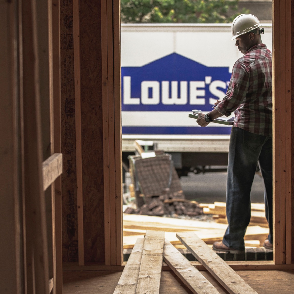 Image d'un ouvrier de la construction regardant une planchette à pince au milieu de l’image et d'un camion Lowe's en arrière-plan